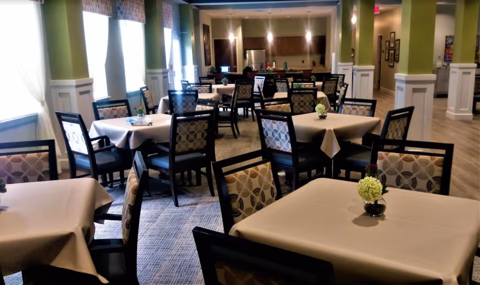 A dining room with multiple square tables covered with beige tablecloths, each surrounded by patterned cushioned chairs. Small floral centerpieces are placed on each table. The room has green walls with white wainscoting, large windows with sheer curtains, and a kitchen area with cabinets and a refrigerator in the background.