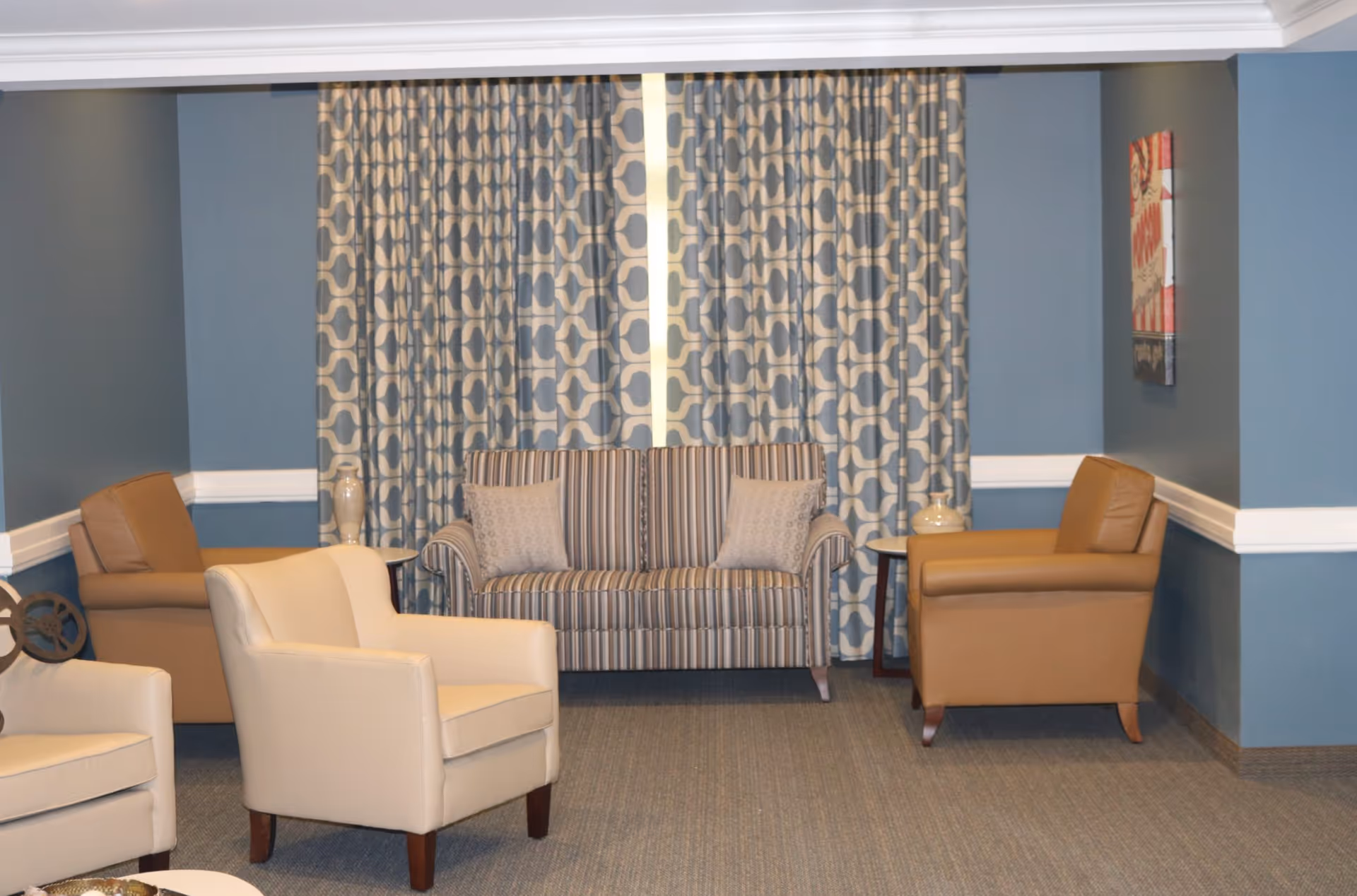 A cozy seating area in a senior living facility featuring a striped loveseat with two patterned pillows, two tan leather armchairs, and two cream-colored armchairs arranged on a carpeted floor. The background has blue walls with white trim and patterned curtains covering a window. There is a small side table with a decorative vase next to each leather armchair and a piece of abstract wall art on the right wall.