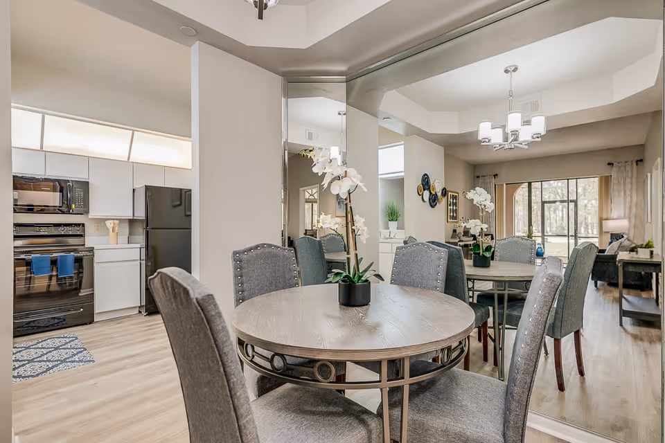 A round dining table with upholstered chairs and orchid centerpieces in a mirrored dining area opening to a kitchen and living room.