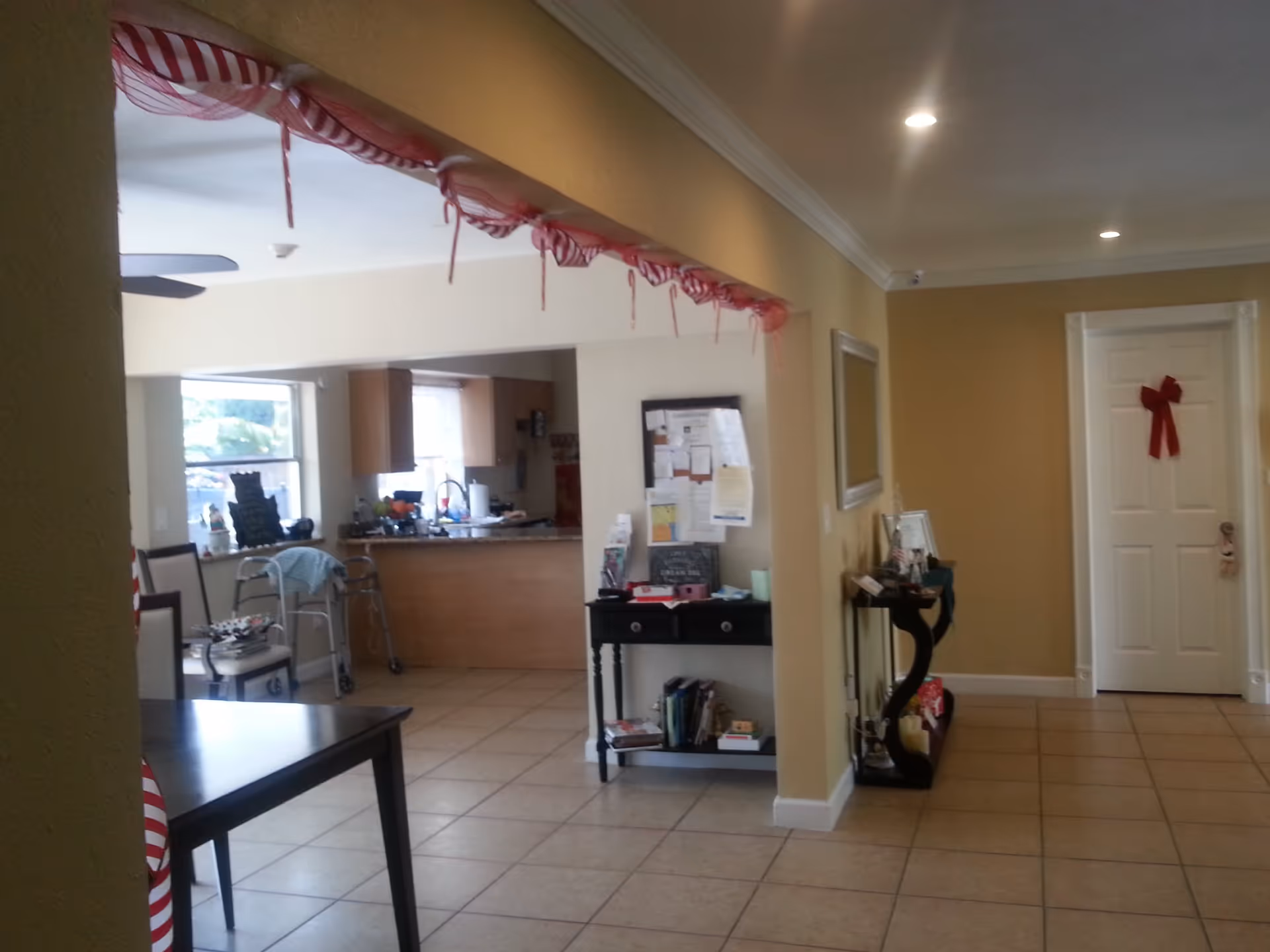 Open interior common area with tiled floor showing a kitchen pass-through, table, walker, bulletin board, and a door decorated with a red bow.