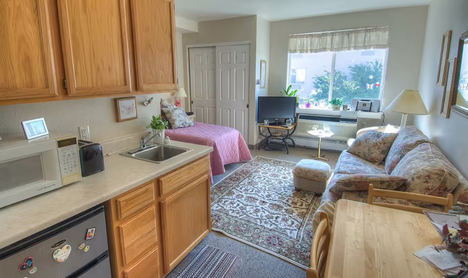 A cozy assisted living studio apartment featuring a small kitchenette with wooden cabinets, a microwave, and a mini refrigerator. The room includes a single bed with a pink quilt, a floral-patterned couch with matching pillows, a small ottoman, a wooden dining table with chairs, and a TV on a stand near a large window with plants on the windowsill. The space is well-lit with natural light coming through the window.