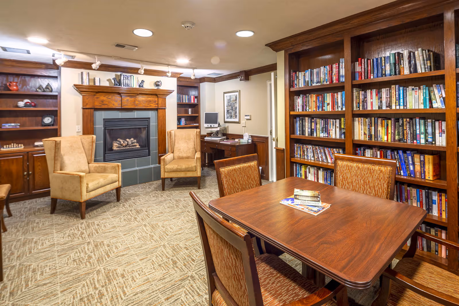 Cozy senior living library lounge with bookshelves, a fireplace, a table and chairs.