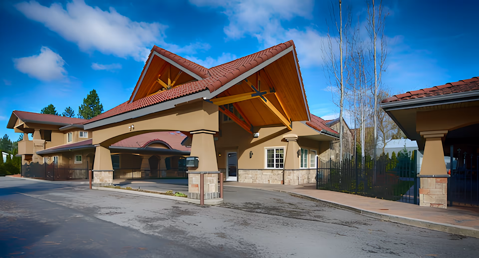 Exterior view of a senior living facility building with a covered entrance supported by large columns, beige walls, and a red tiled roof under a blue sky with some clouds.