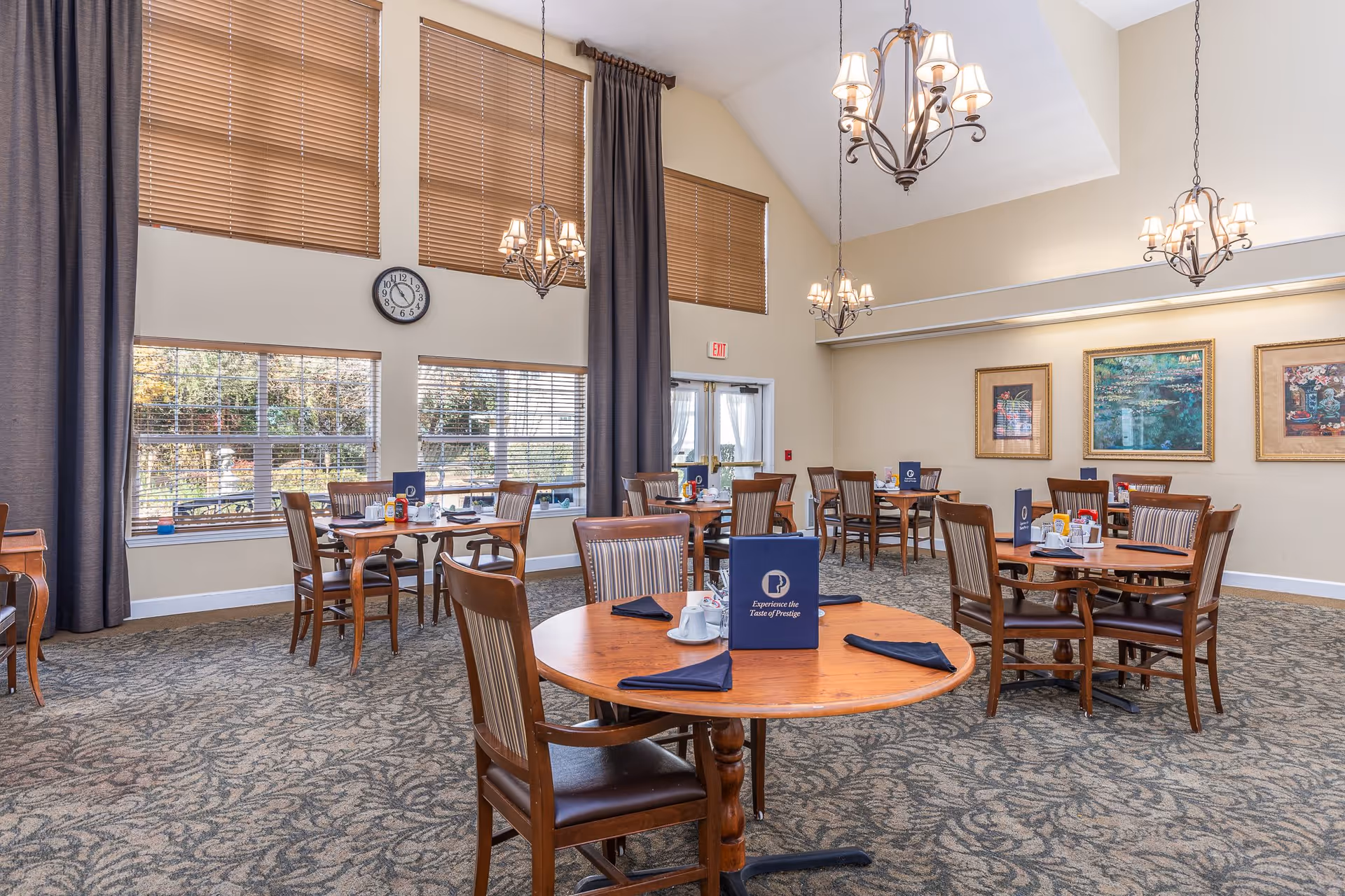 A bright and spacious dining room with several round wooden tables and chairs arranged neatly. Each table has a blue menu, napkins, and condiments. Large windows with brown blinds and tall gray curtains let in natural light. The room features beige walls adorned with framed artwork and three elegant chandeliers hanging from the ceiling. A clock is mounted on the wall above the windows.