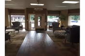 A senior living facility lobby with upholstered chairs and small tables arranged on a wood floor facing glass entrance doors and windows.