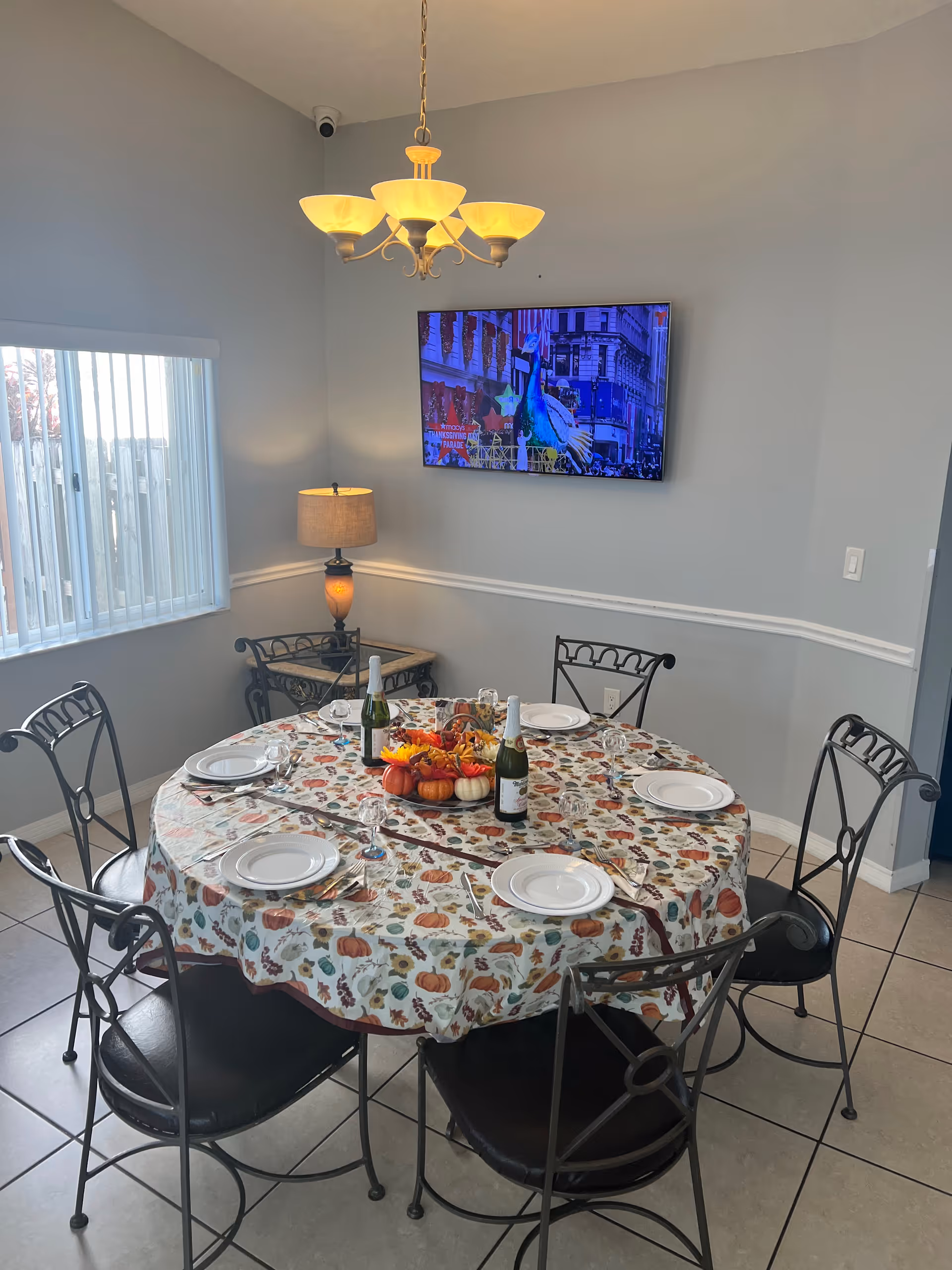 Round dining table set for six with a fall-themed tablecloth and centerpiece under a chandelier in a bright dining room with a wall-mounted TV.