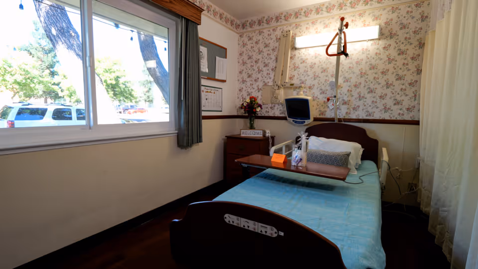 A patient bedroom with a hospital bed, overbed table, monitor, floral wallpaper and a window looking out to parked cars.