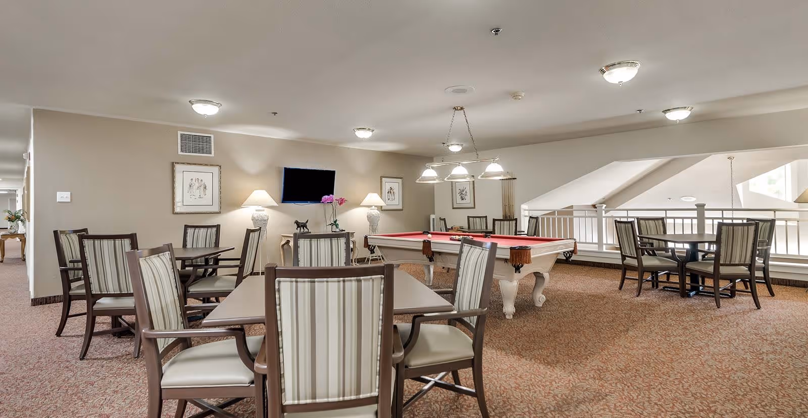A spacious common area with multiple tables and chairs arranged for seating. In the center, there is a white pool table with a red felt top and a hanging light fixture above it. The room has beige walls, carpeted floors, framed artwork on the walls, and several ceiling lights. A flat-screen TV is mounted on one wall with two table lamps on either side.
