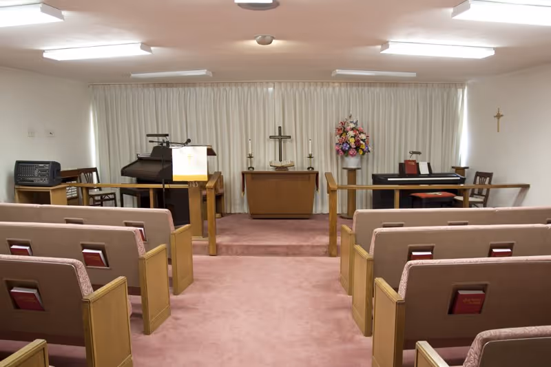Small chapel interior with rows of pews facing an altar, a cross, piano/organ, and a floral arrangement.
