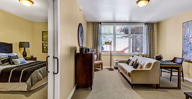Bright suite featuring a living area with a sofa and desk by a large window, and an adjacent bedroom visible through a sliding door.