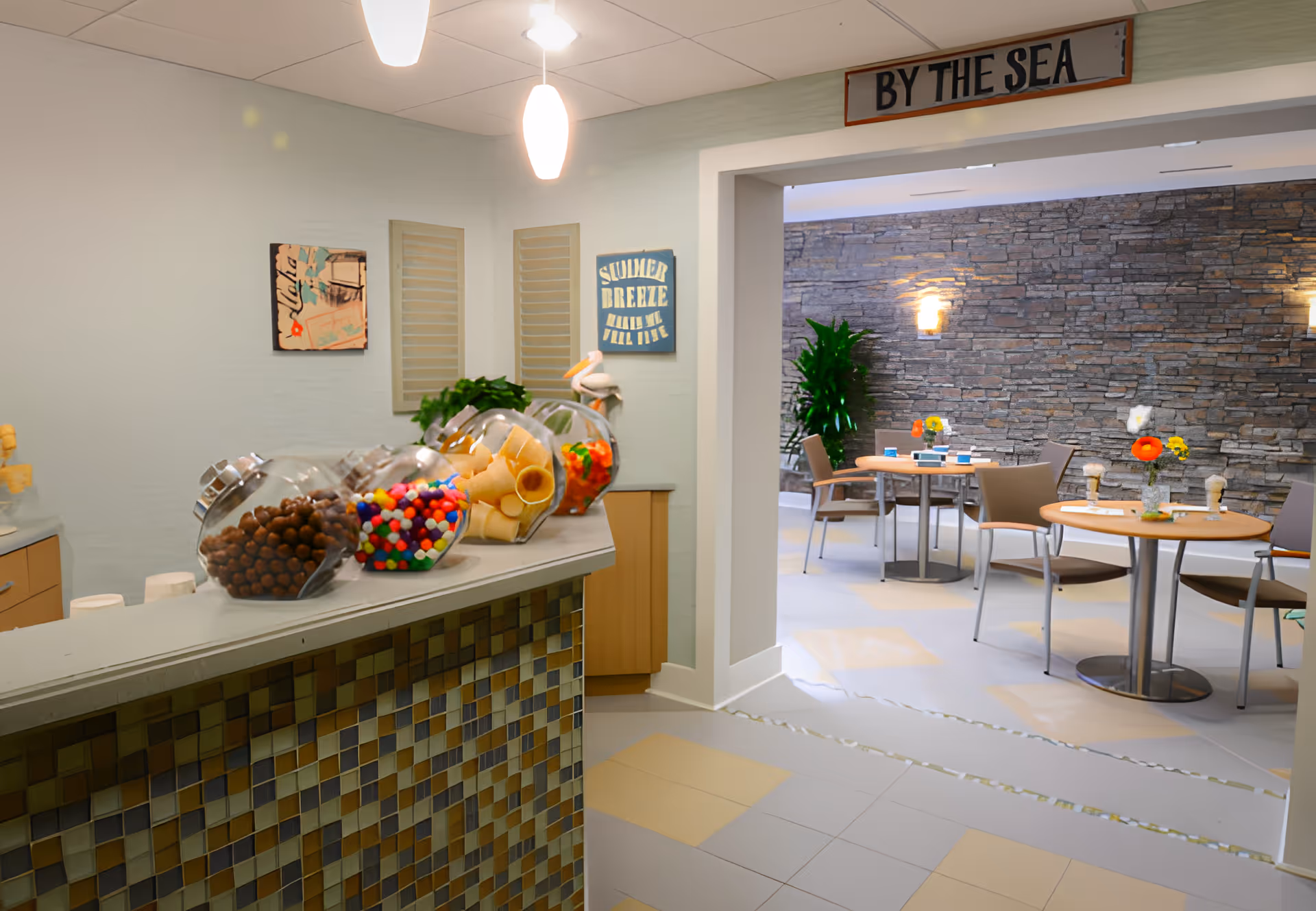 Interior view of a dining area in a senior living facility featuring a counter with jars of snacks and a seating area with round tables and chairs. The walls have decorative signs, including one that says 'BY THE SEA'. The space is well-lit with modern lighting and has a stone accent wall in the background.