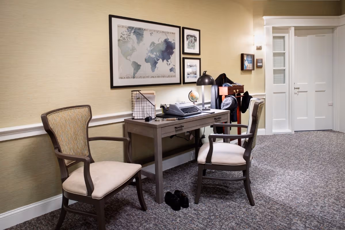 A cozy interior corner featuring a wooden desk with a typewriter, a small globe, a desk lamp, and some papers. Two cushioned wooden chairs are placed on either side of the desk. The wall behind the desk has framed artwork, including a world map and smaller pictures. A coat rack with hats and coats is visible next to the desk, and a pair of black shoes is on the carpeted floor. The room has beige walls and a white door in the background.