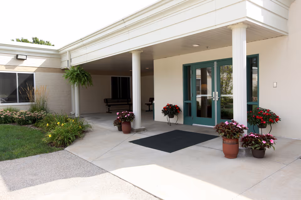 Covered entrance with white columns, green double doors, a doormat, and potted flowers.