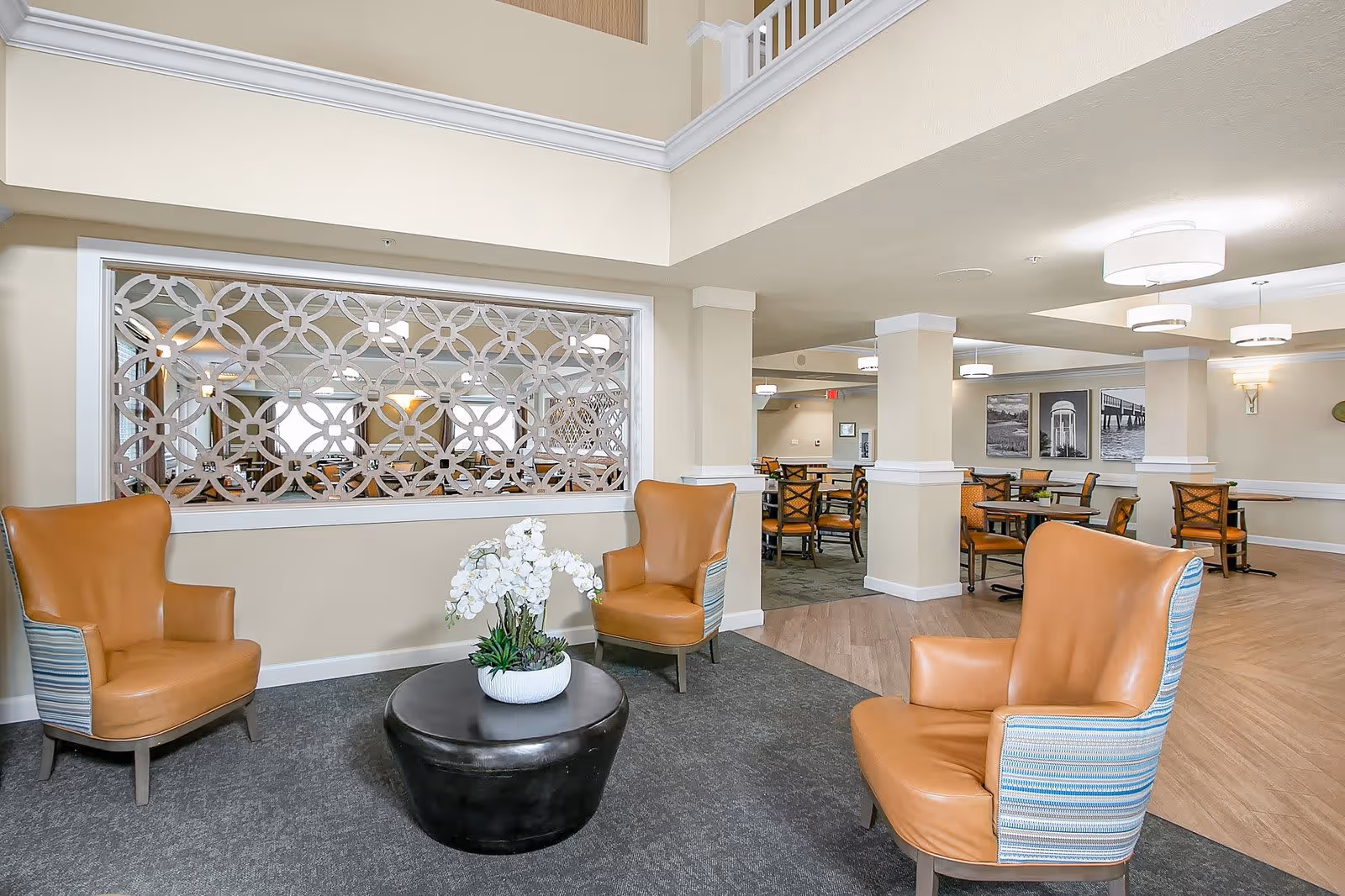 A seating area with three tan leather armchairs arranged around a round black coffee table with a white flower arrangement. Behind the seating area is a decorative partition with a geometric pattern, and beyond that is a dining area with tables and chairs. The room has beige walls, white trim, and modern ceiling lights.