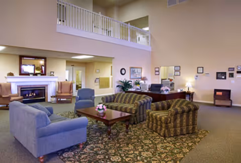 Spacious senior living facility common area with high ceilings and a balcony railing above. The room features a fireplace with two armchairs nearby, a blue sofa, two patterned armchairs, and a wooden coffee table with a small flower arrangement. There is a patterned area rug under the seating area, and the walls are decorated with framed pictures and a clock. The space is well-lit with ceiling lights and has a warm, inviting atmosphere.