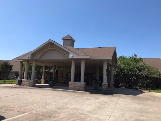 Front exterior view of a single-story building with a covered entrance supported by columns, surrounded by some trees and a clear blue sky.