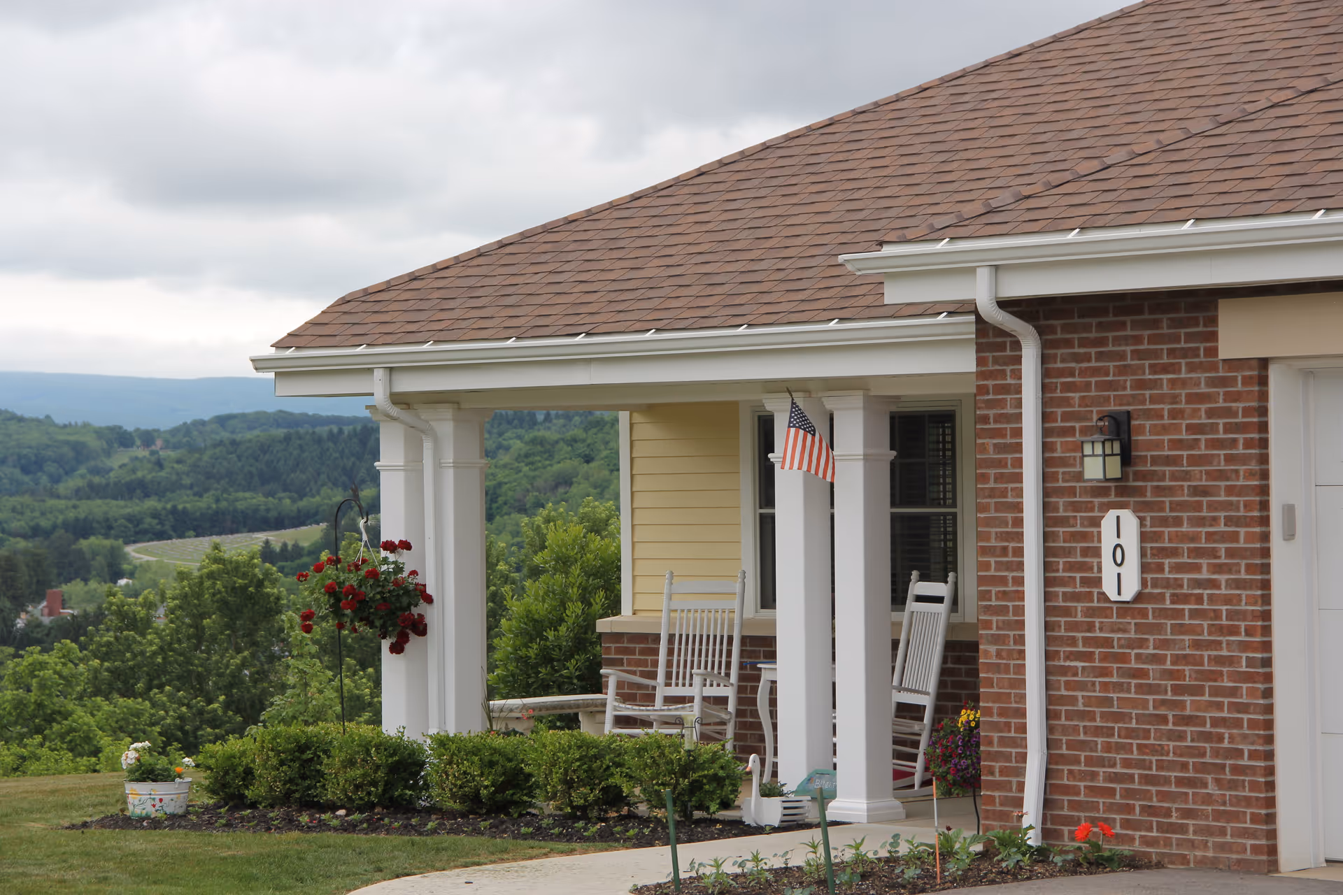 Front porch of a residential building with two white rocking chairs, a small table, hanging red flowers, and an American flag attached to a white column. The building has a brick and yellow siding exterior with a brown shingled roof. There is a house number plaque displaying 101 and a scenic view of green trees and hills in the background under a cloudy sky.