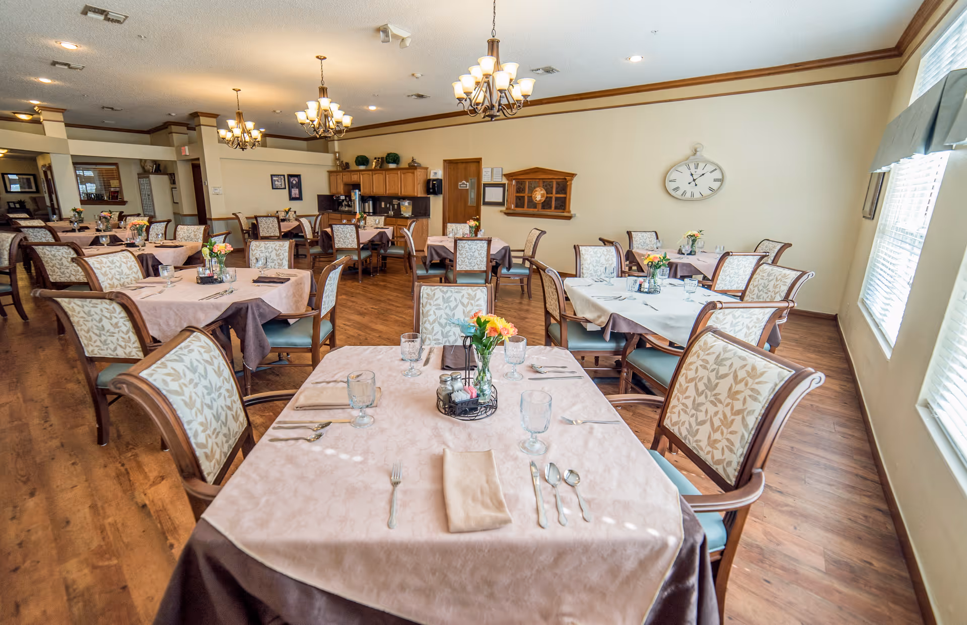 Dining room with neatly set tables and upholstered chairs, chandeliers, wood floors, and large windows.