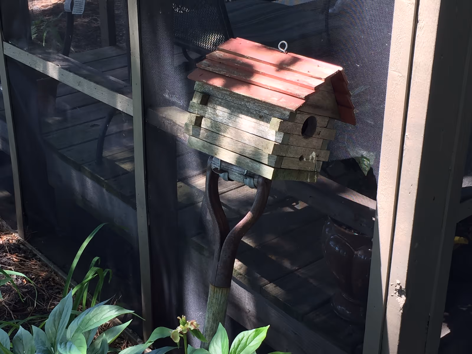 Wooden birdhouse with a red roof mounted on a garden tool in front of a screened porch and surrounding plants.