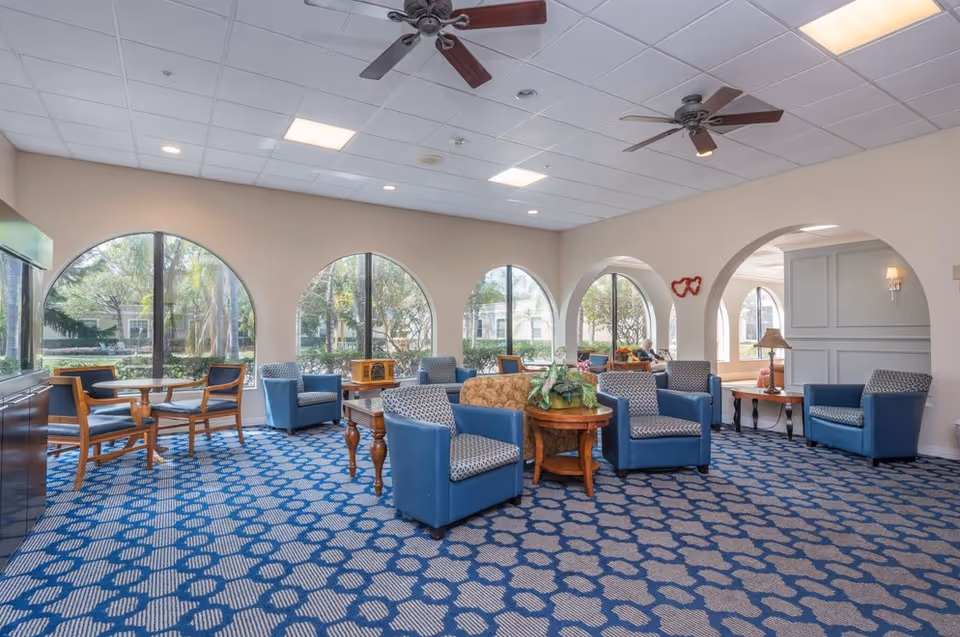 A spacious living room area with patterned blue carpet, multiple blue and patterned armchairs, wooden tables with plants, large arched windows letting in natural light, ceiling fans, and beige walls with archways leading to another room.