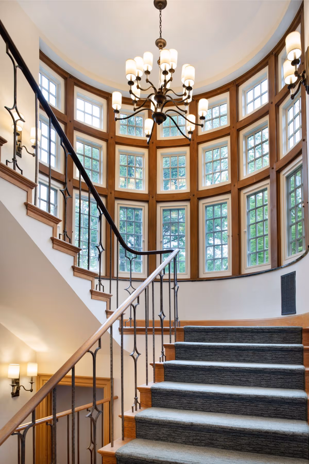 Carpeted staircase with wooden handrails and decorative iron balusters leading up to a curved wall of large windows with wooden frames, illuminated by a central chandelier and wall sconces.