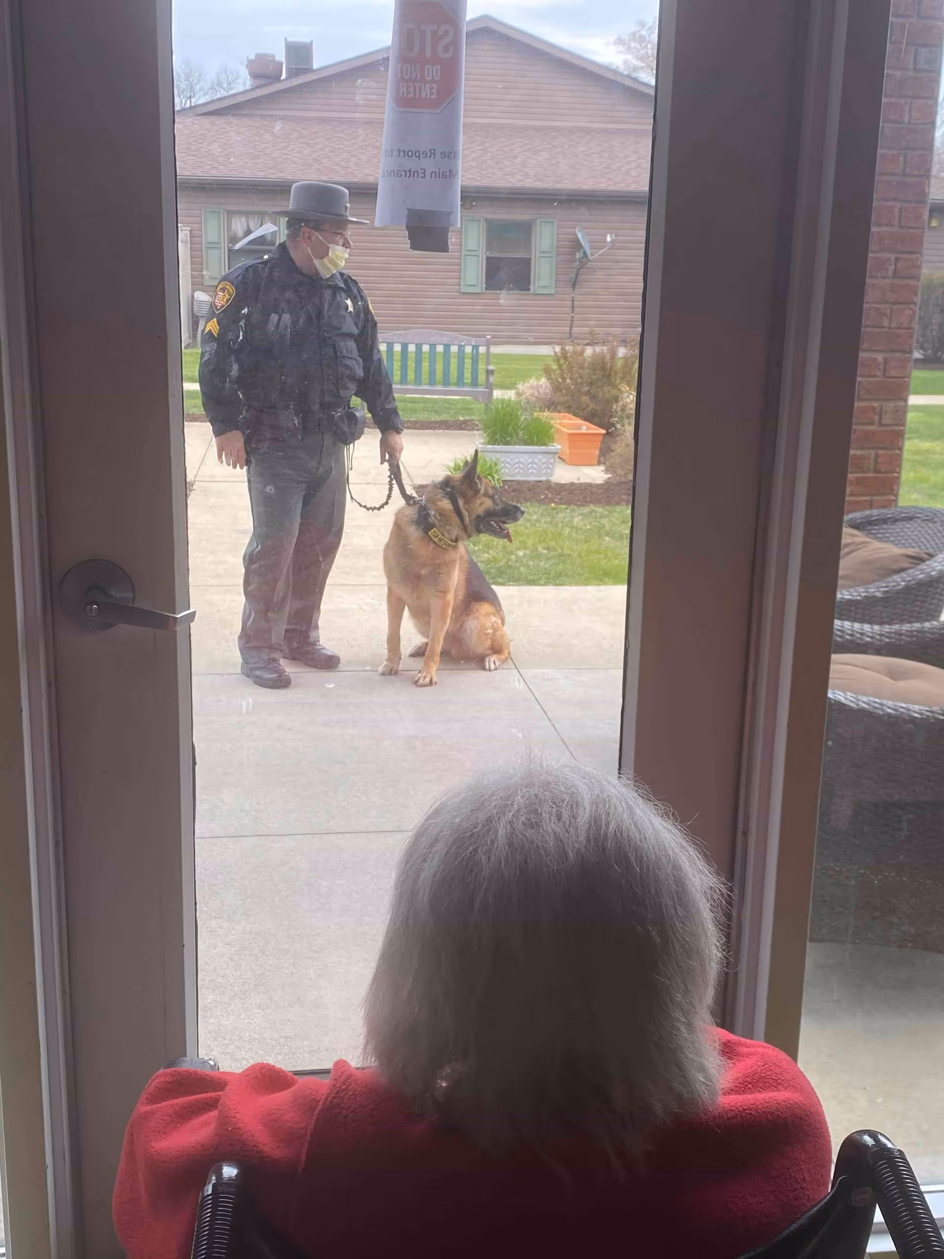 An elderly person with white hair wearing a red jacket sits in a wheelchair indoors, looking out through a glass door at a uniformed police officer holding a leash attached to a German Shepherd dog standing outside on a sidewalk. A house and garden with plants and a bench are visible in the background.