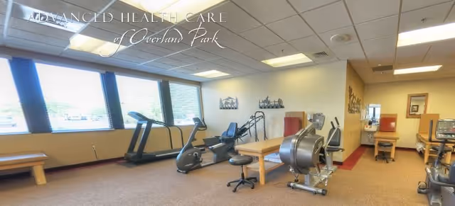 A fitness room in Advanced Health Care of Overland Park featuring exercise equipment including a treadmill, stationary bike, rowing machine, and other workout apparatus. The room has large windows with blinds, beige walls, and a carpeted floor. There are benches and stools placed around the equipment.