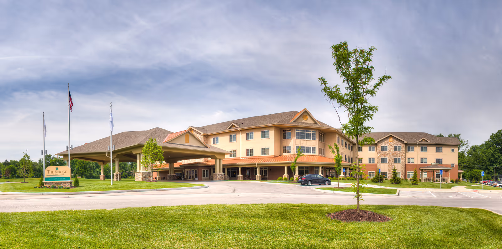 Exterior view of a large, multi-story senior living facility building with a covered entrance driveway, surrounded by green lawns and trees under a partly cloudy sky.