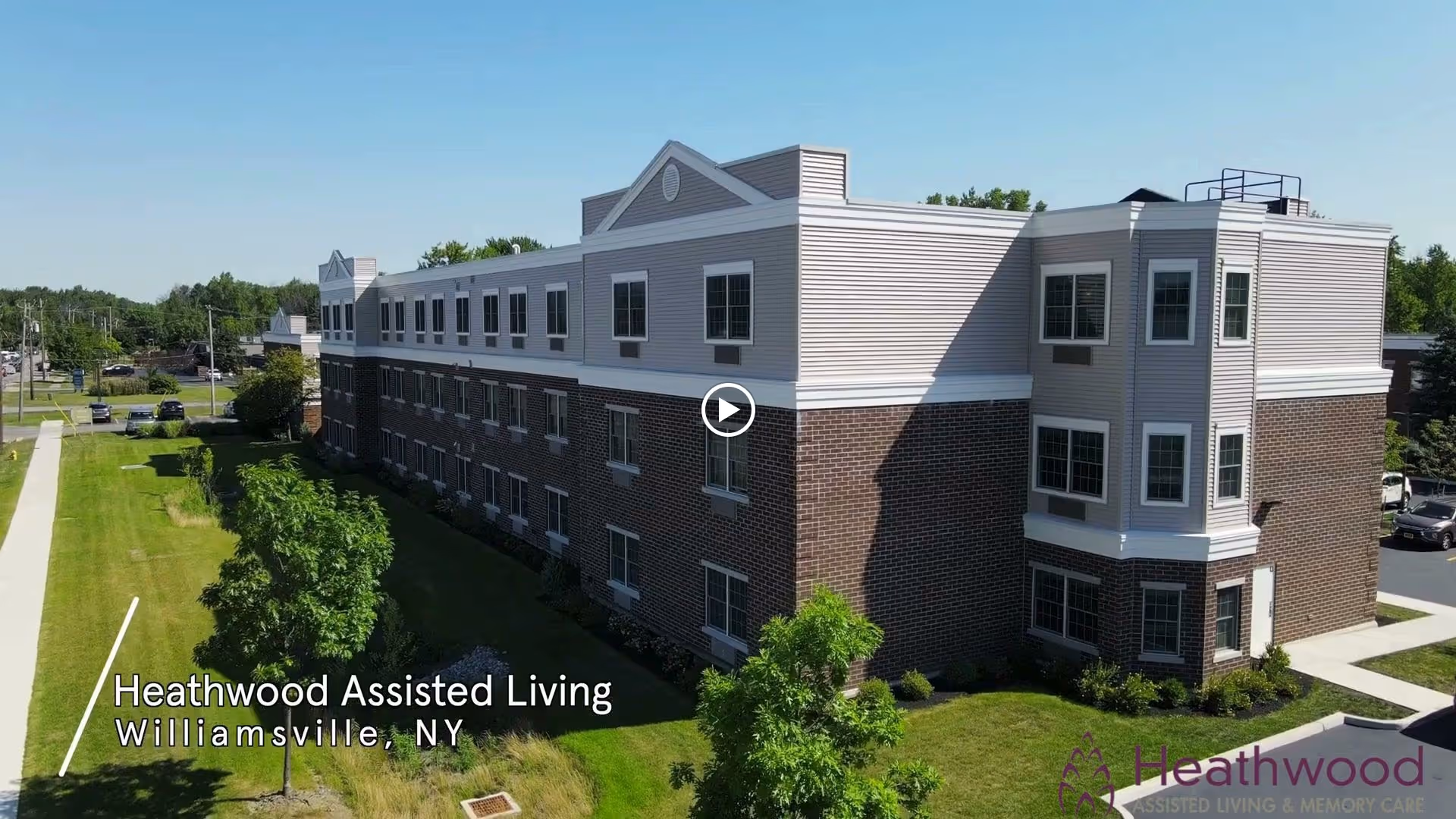 Exterior view of a three-story assisted living facility building with brick and light-colored siding, surrounded by green grass and trees under a clear blue sky. The building has multiple windows and a small parking area visible on the right side.