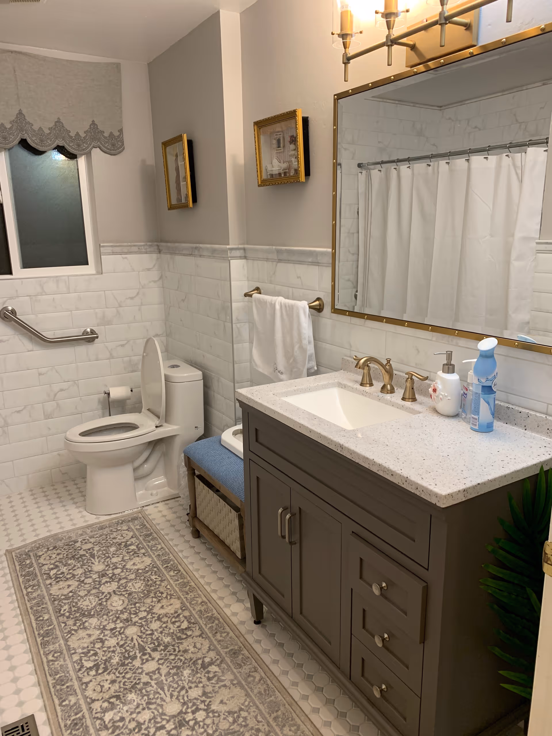 A clean and well-lit bathroom featuring a white toilet with the lid open, a gray vanity with a white countertop and gold fixtures, a large mirror with gold trim, a towel hanging on a gold towel rack, a decorative rug on the tiled floor, a window with a decorative valance, and a shower area with a white curtain.