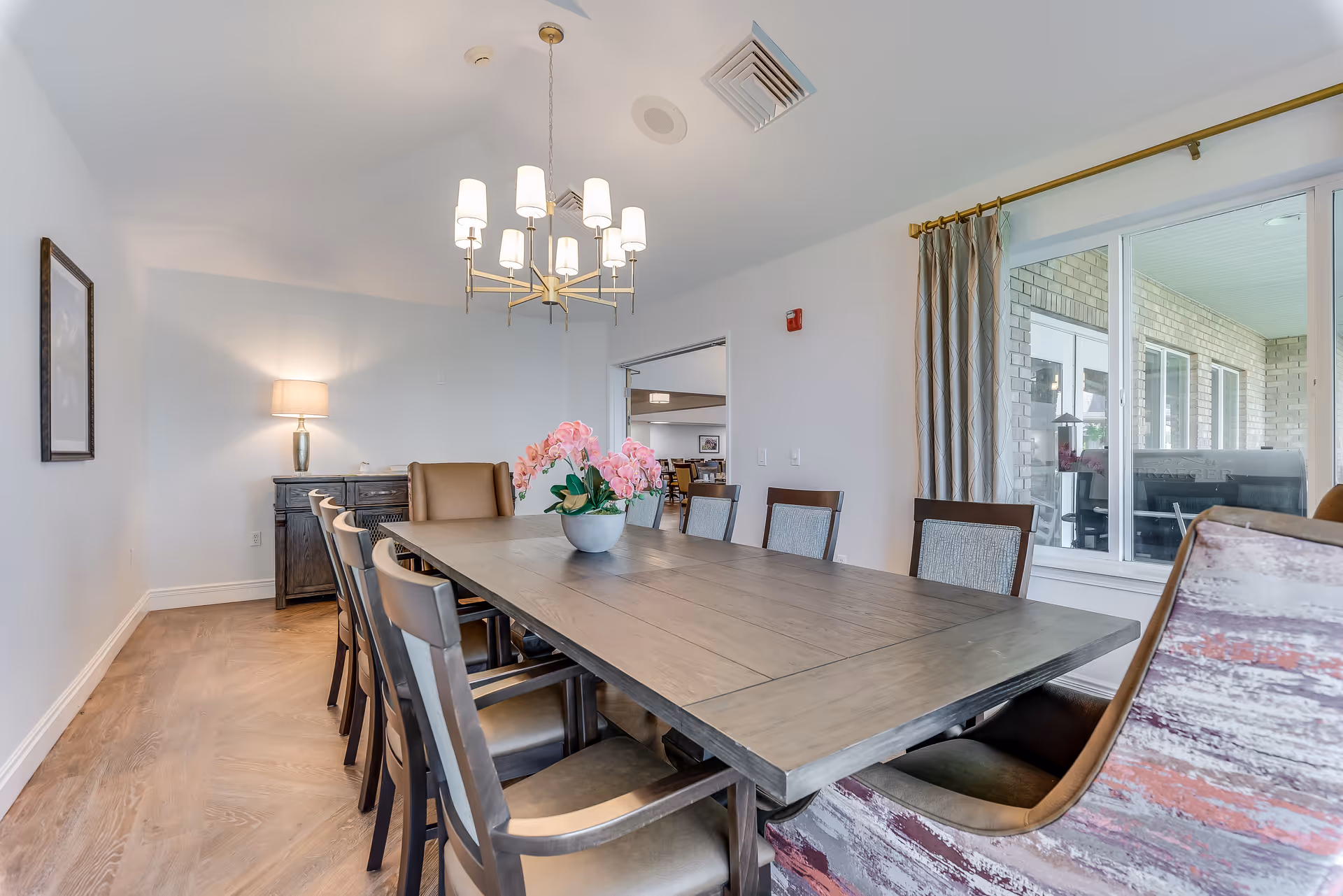 Bright dining room with a long wooden table surrounded by chairs, a chandelier overhead, and a vase of pink flowers near windows overlooking a patio.