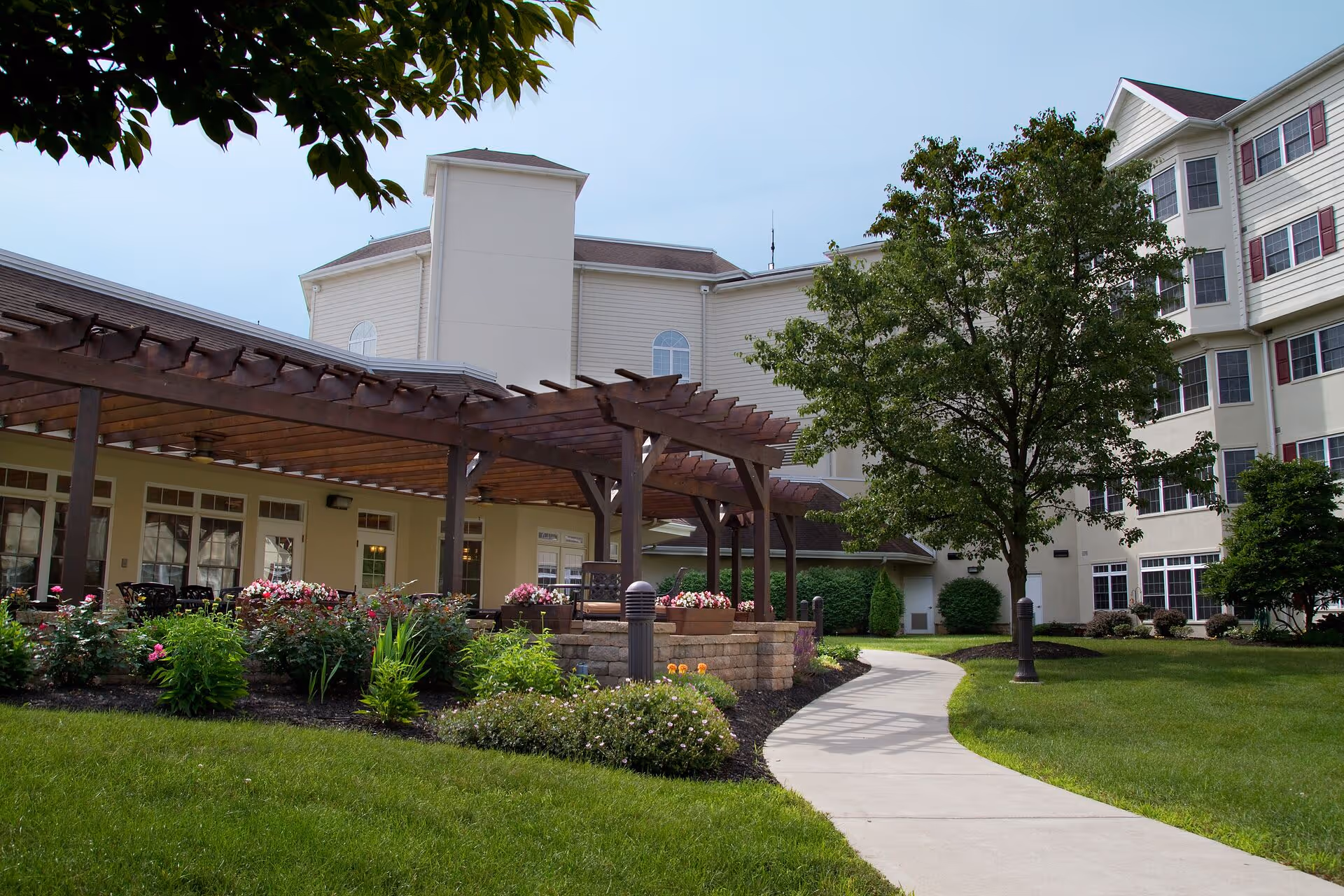 Outdoor courtyard and walkway beside a multi-story senior living building with a wooden pergola, landscaping, and seating.