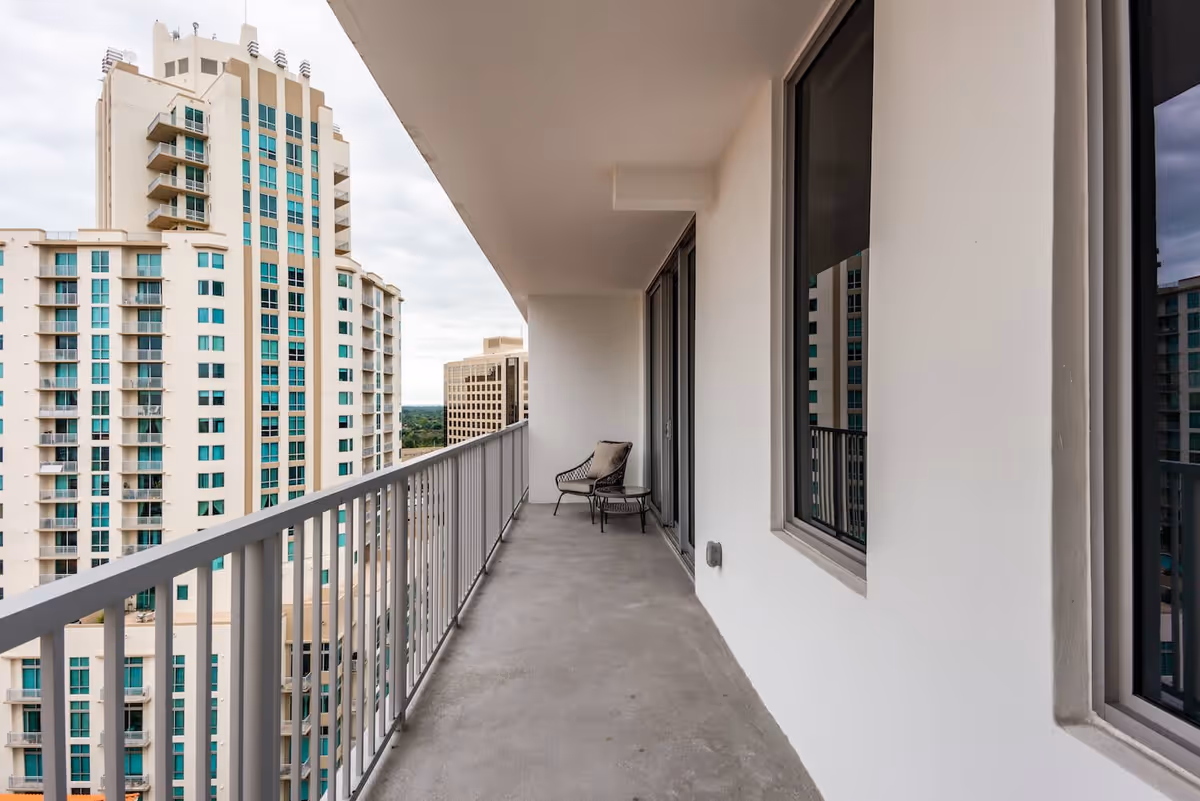 A long balcony with a metal railing on the left side and two chairs with a small table at the far end. The balcony overlooks tall buildings with multiple windows and balconies under a cloudy sky.