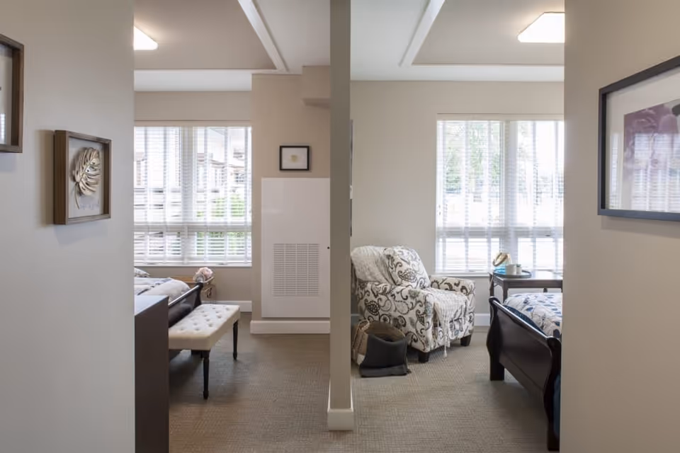 Interior view of a senior living facility bedroom area with two separate sleeping spaces divided by a partial wall. Each space has a bed, a window with blinds, and seating. One side features a cushioned bench at the foot of the bed, and the other side has a patterned armchair with a blanket draped over it and a small side table with a hat and a tissue box.