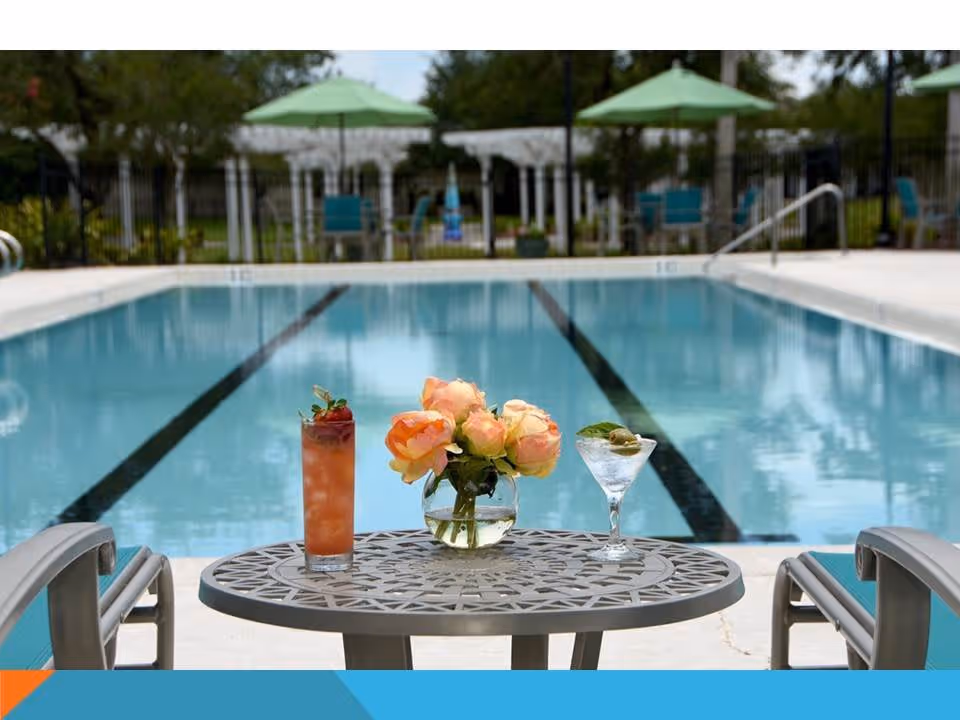A poolside scene featuring a round metal table with a vase of peach-colored roses and two drinks, one a cocktail with olives and the other a tall iced beverage. The swimming pool with lane markings is in the background, along with green umbrellas and chairs on the far side of the pool.