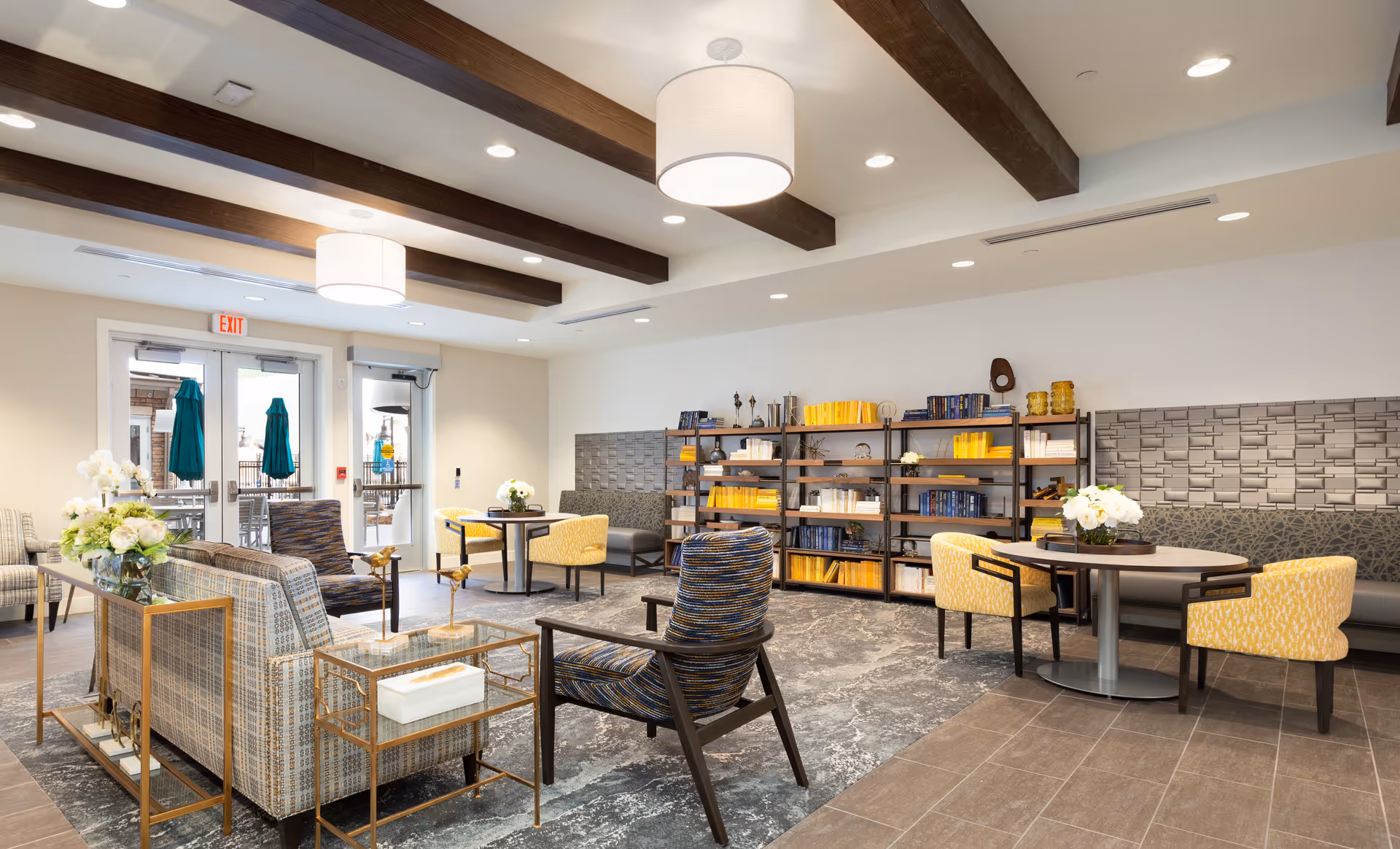 A bright, modern communal lounge with sofas, armchairs, tables and bookshelves under exposed ceiling beams.