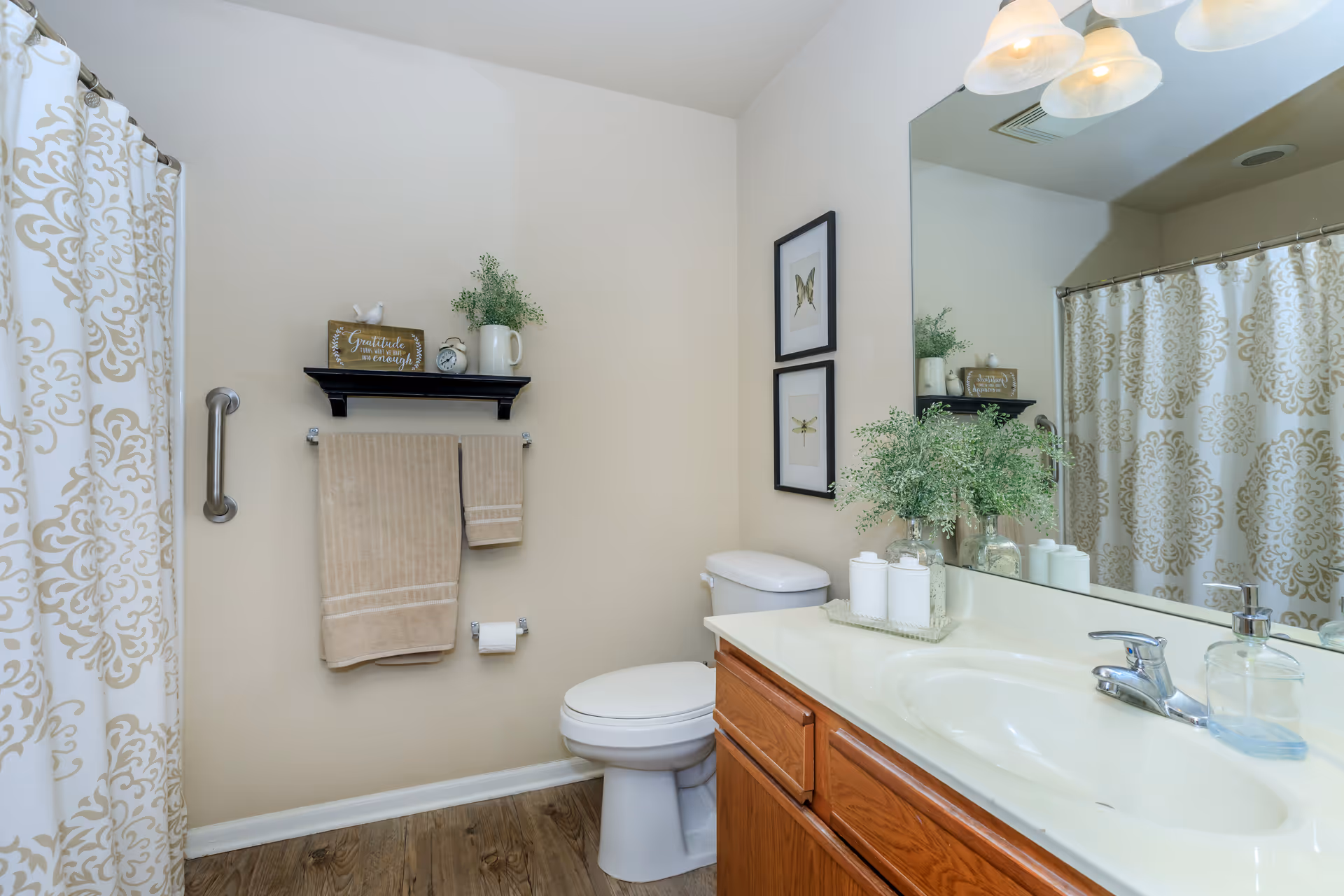A clean and well-lit bathroom featuring a white toilet, a wooden vanity with a white countertop and sink, and a large mirror above the sink. There is a decorative plant and containers on the countertop. Beige towels hang on a towel rack beneath a small black shelf holding a small plant, a clock, and a sign that reads 'Gratitude is enough.' The shower curtain has a beige and white ornate pattern, and the floor is wood-style.