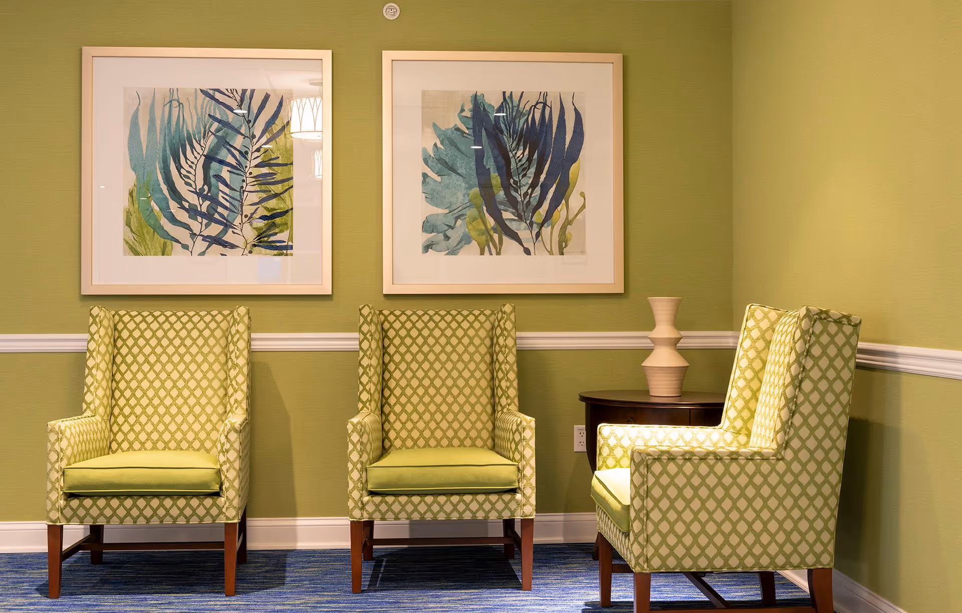 Three green patterned armchairs arranged in a corner of a room with green walls and blue carpet. Two framed botanical prints hang on the wall above the chairs, and a small dark wooden side table with a beige decorative vase is placed between two of the chairs.