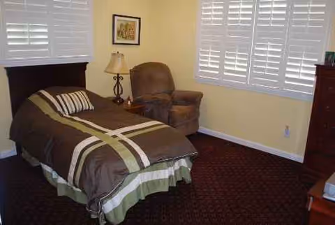 A furnished bedroom with a bed, chair, lamp, dresser, and large windows with blinds.