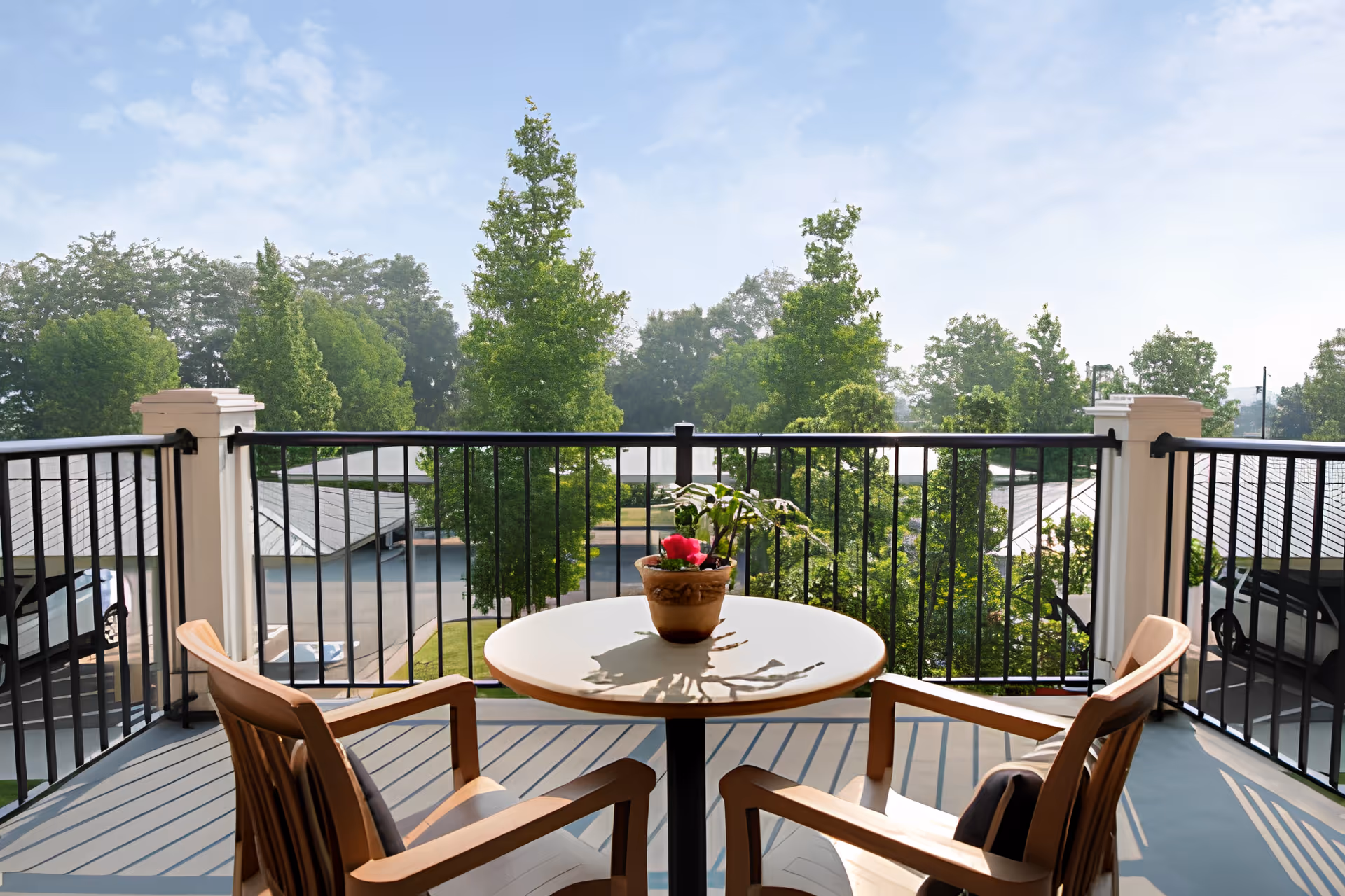 A balcony with a round table and two wooden chairs facing outward. On the table is a potted plant with a pink flower. The balcony has a black metal railing and overlooks a parking area and lush green trees under a partly cloudy sky.