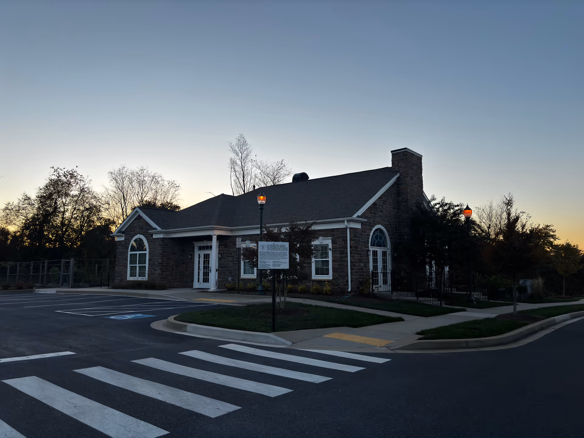 Exterior view of a single-story stone building with large arched windows and a chimney, surrounded by a parking lot with marked spaces and a pedestrian crosswalk, taken at dusk with street lamps lit and trees in the background.