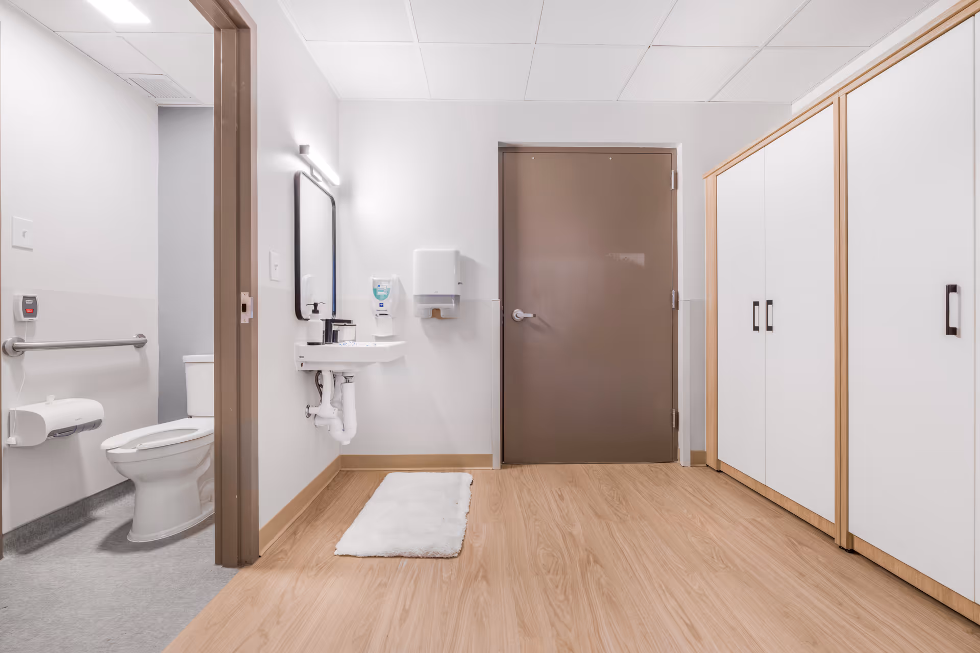 Interior view of a clean and modern bathroom area in a healthcare facility. The image shows a toilet with a grab bar, a sink with a mirror above it, a soap dispenser, a paper towel dispenser, a brown door, and white cabinets with wooden trim. The floor is light wood laminate, and there is a white rug in front of the sink.
