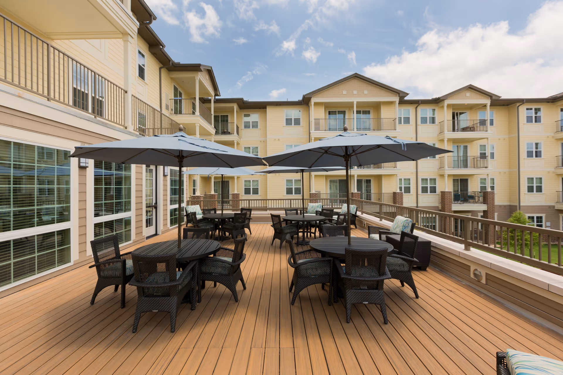 Outdoor patio area at The Enclave at Cedar Park Senior Living with multiple round tables, black wicker chairs, and large gray umbrellas. The patio is surrounded by a railing and overlooks a multi-story yellow building with balconies and windows under a partly cloudy sky.