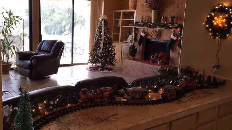 A cozy living room decorated for Christmas with a small decorated Christmas tree near a sliding glass door, a fireplace with stockings hanging on the mantel, and a miniature train set with lights on a tiled counter in the foreground. There is a comfortable armchair near the door and a wreath with lights on the wall.