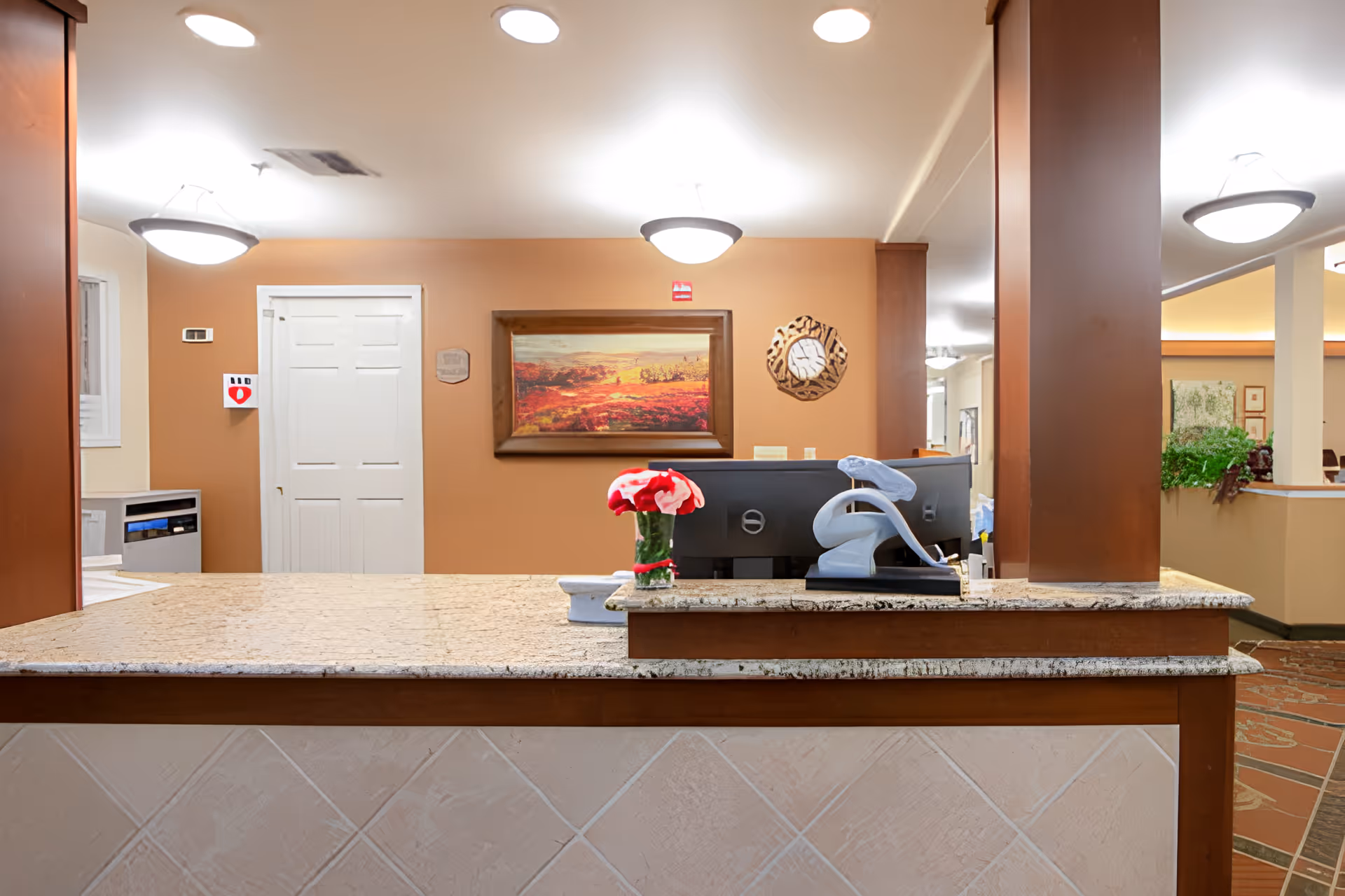 Reception desk in a senior living facility lobby with a granite countertop, computer monitor, vase of flowers, wall art and warm lighting.