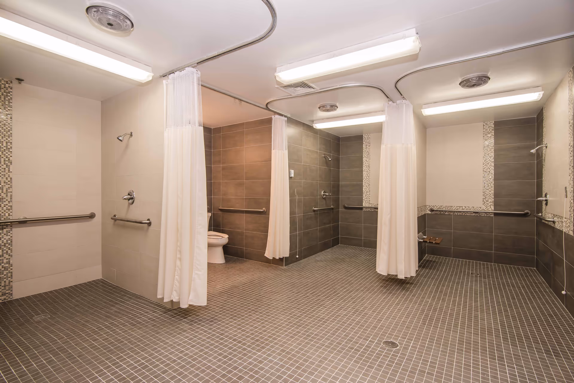 A spacious, accessible bathroom area with multiple shower stalls separated by white curtains. The walls are tiled in neutral tones with grab bars installed for safety. There is a toilet visible in one of the stalls, and the floor is covered with small square tiles. The ceiling has fluorescent lighting and ventilation fixtures.