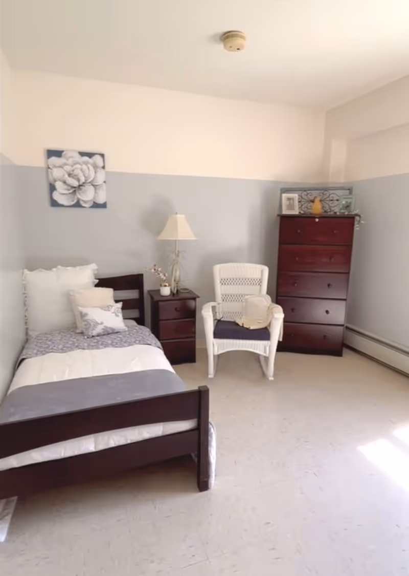 A simple bedroom with a single dark wooden bed with white and gray bedding, a matching dark wooden nightstand with a lamp and small plant, a white wicker chair with a cushion and a light-colored bag on it, and a tall dark wooden dresser with decorative items on top. The walls are painted light gray on the bottom half and white on the top half, with a flower artwork hanging above the bed.