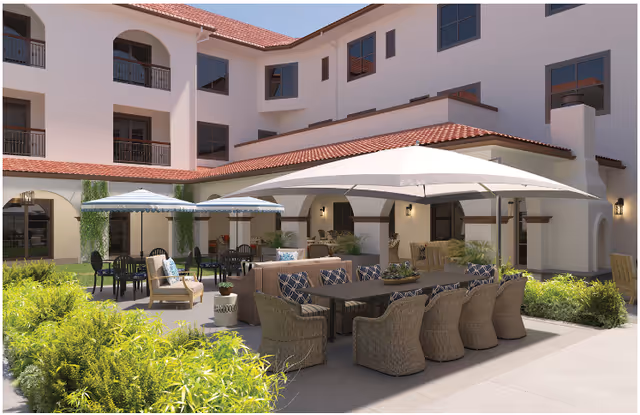 Outdoor courtyard area of The Hacienda Mission San Luis Rey featuring multiple seating arrangements including wicker chairs with cushions around a long table under a large white umbrella, additional tables with blue umbrellas, and greenery surrounding the paved patio. The building has white walls, red tile roofing, and multiple windows and balconies.