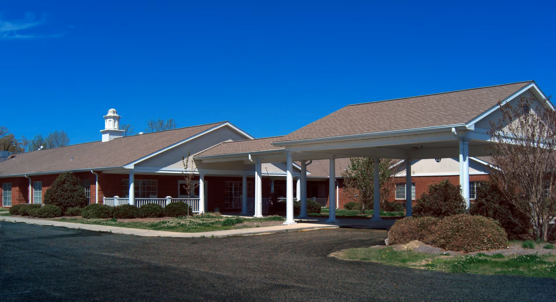 Single-story brick senior living facility with a covered drop-off portico, white columns, and landscaping under a clear blue sky.