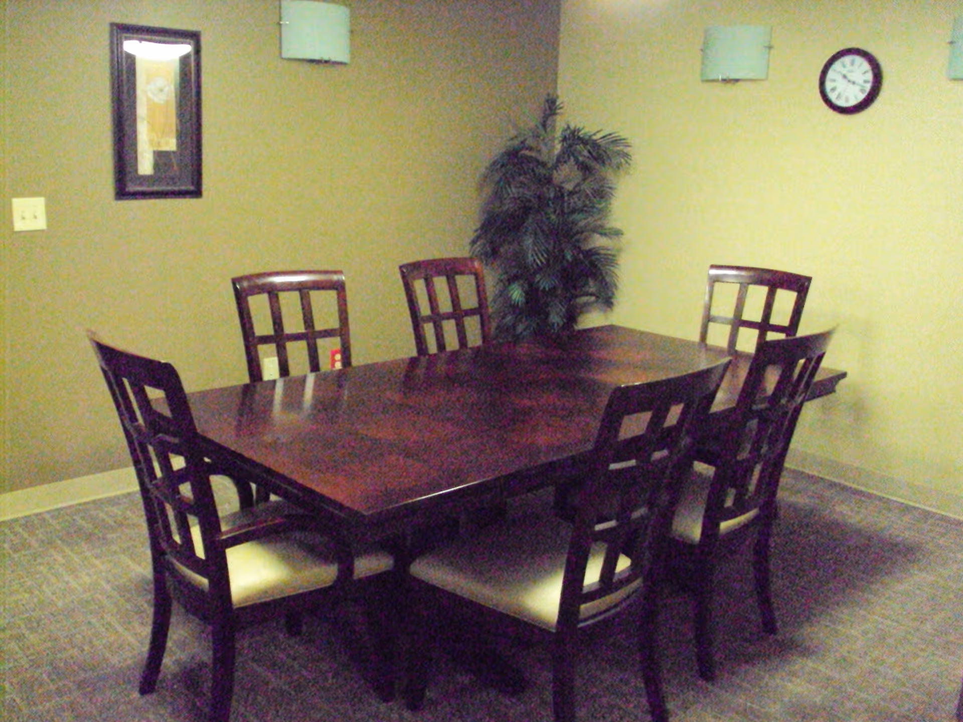 A wooden dining table surrounded by six chairs in a small room with a potted plant and a wall clock.